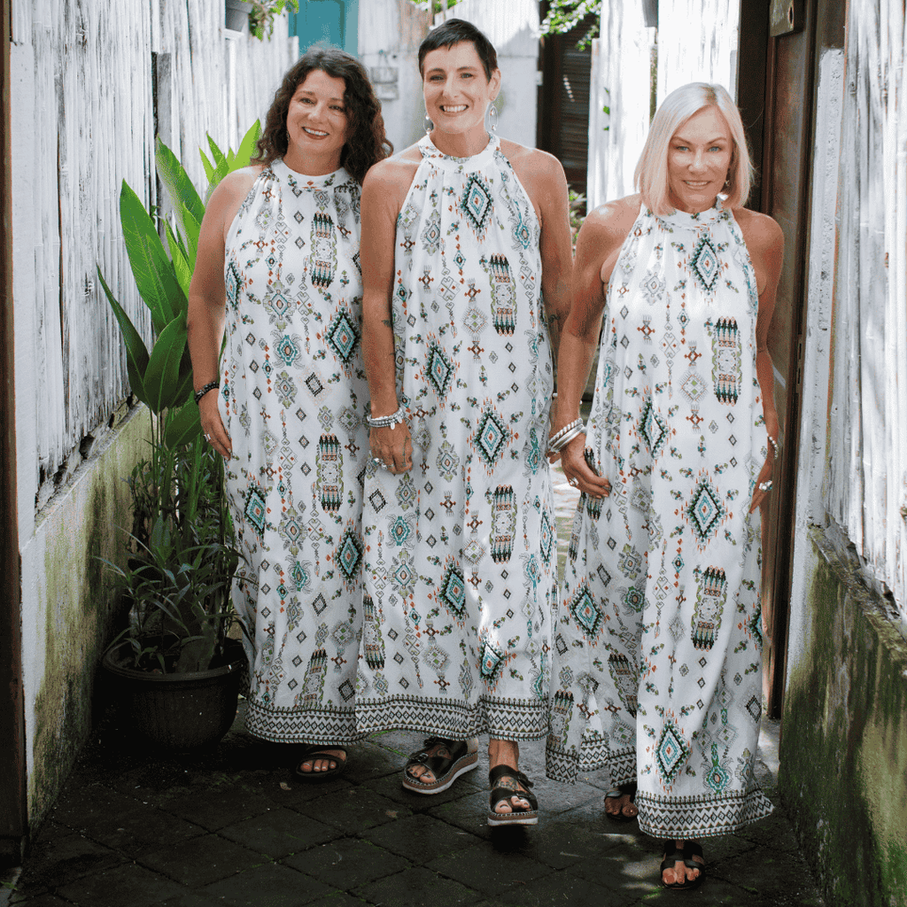 Three menopuasal women wearing matching white dresses with patterns standing together outdoors.