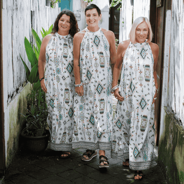 Three menopuasal women wearing matching white dresses with patterns standing together outdoors.